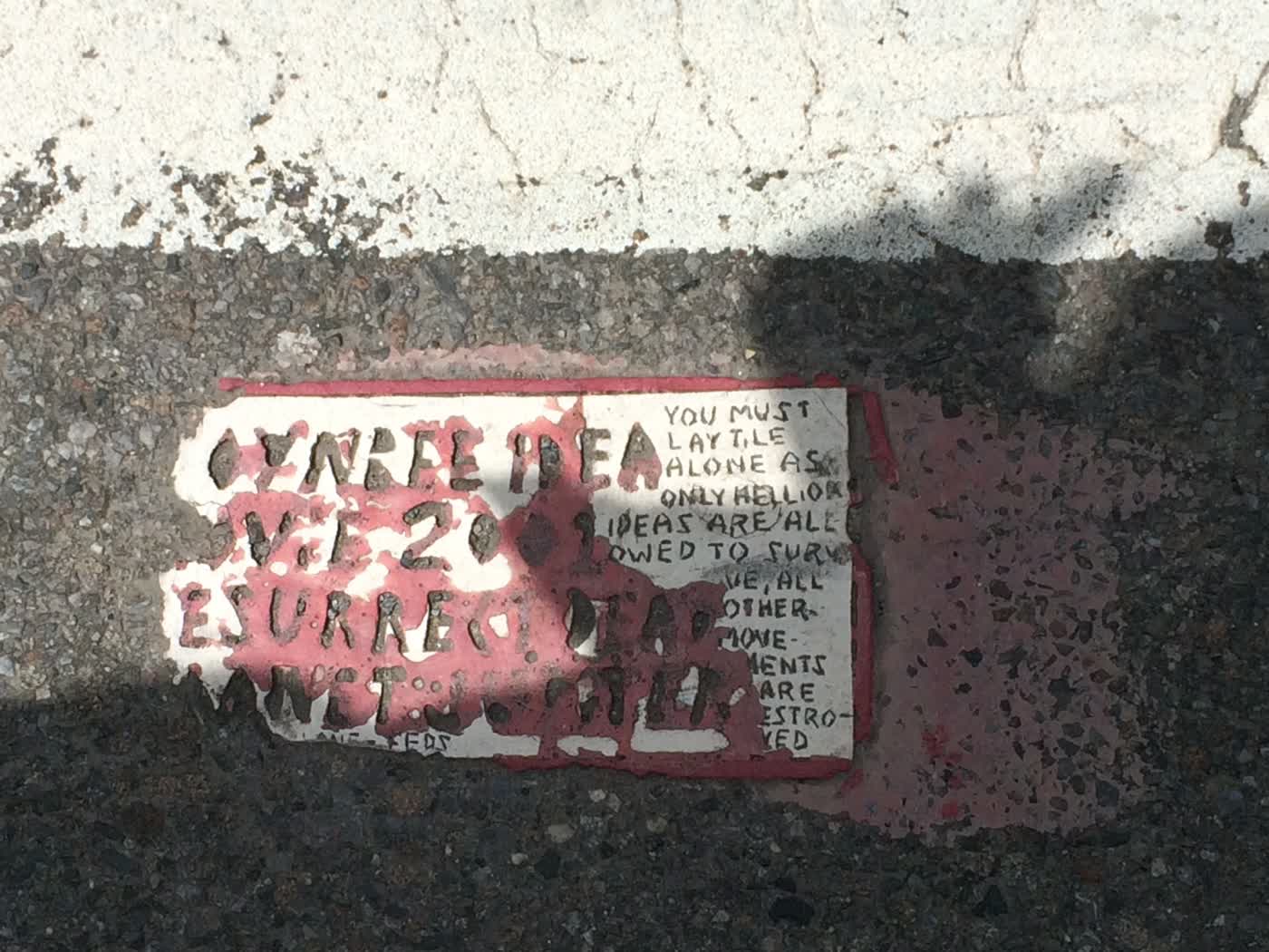 A small tile embedded in a road's asphalt reading, 'TOYNBEE IDEA IN MOVIE 2001, RESURRECT DEAD ON PLANET JUPITER', with a smaller side note reading, 'YOU MUST LAY TILE ALONE AS ONLY HELL OR IDEAS ARE ALLOWED TO SURVIVE, ALL OTHER MOVEMENTS ARE DESTROYED'