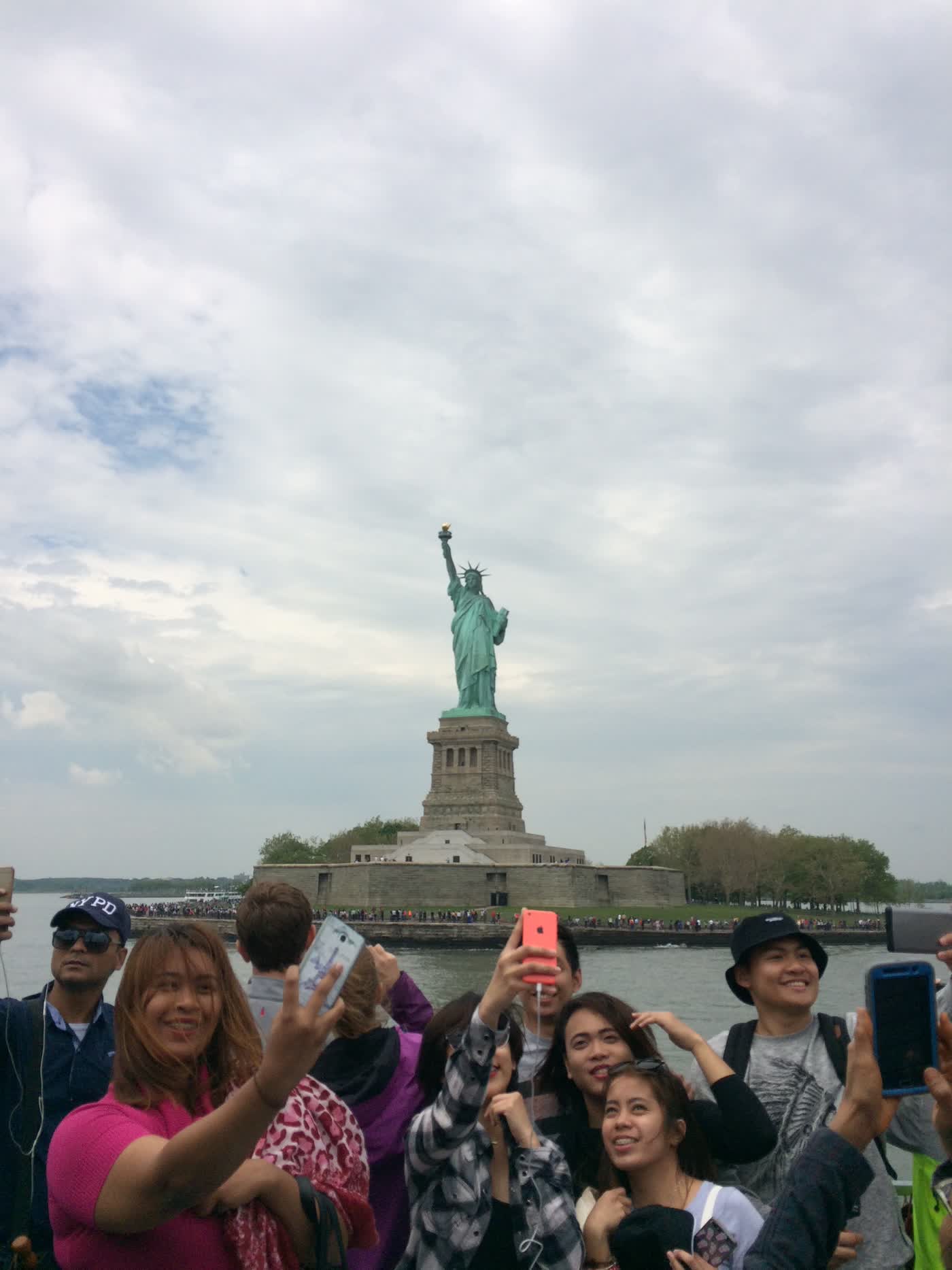 The Statue of Liberty as seen from the ferry, with a crowd of tourists taking selfies in the foreground.